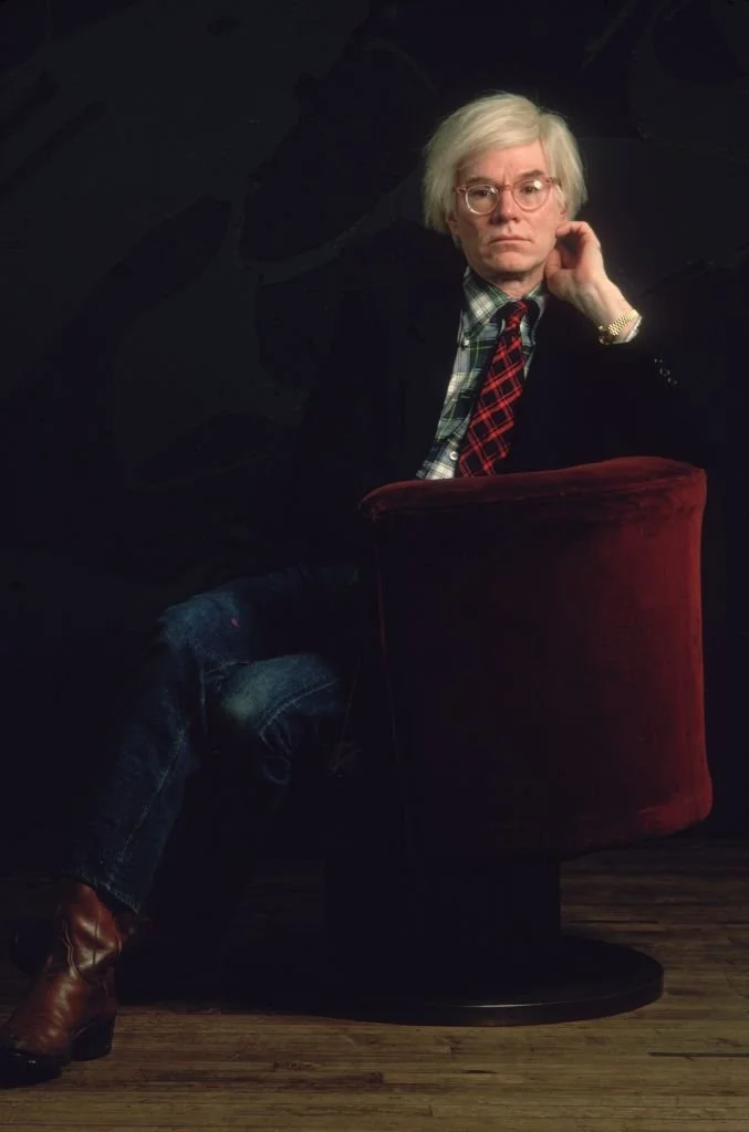 1981: Portrait of American artist Andy Warhol (1928-1987) sitting in a red velvet chair against a black background with his hand up to his face. He wears a jacket and a tie with a plaid shirt, blue jeans and cowboy boots. (Photo by Nancy R. Schiff/Hulton Archive/Getty Images)