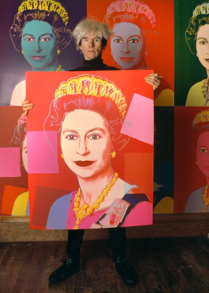 Portrait of American Pop artist Andy Warhol (1928 - 1987) as he holds a silkscreen print of British monarch Queen Elizabeth II in his studio, the Factory, New York, New York, 1985. The picture is part of his Reigning Queens series, others of which are on the wall behind him/Photo by Derek Hudson/Getty Images