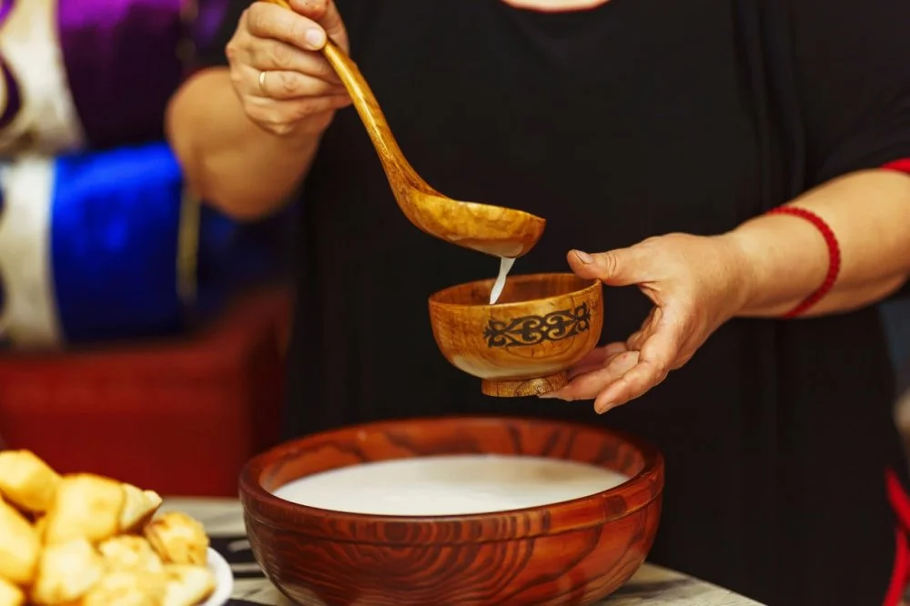 A woman pours "kumis" milk drink into a wooden bowl with an ornament in the national Kazakh style. Central Asian traditional drink on the festive table Nauryz/Shutterstock