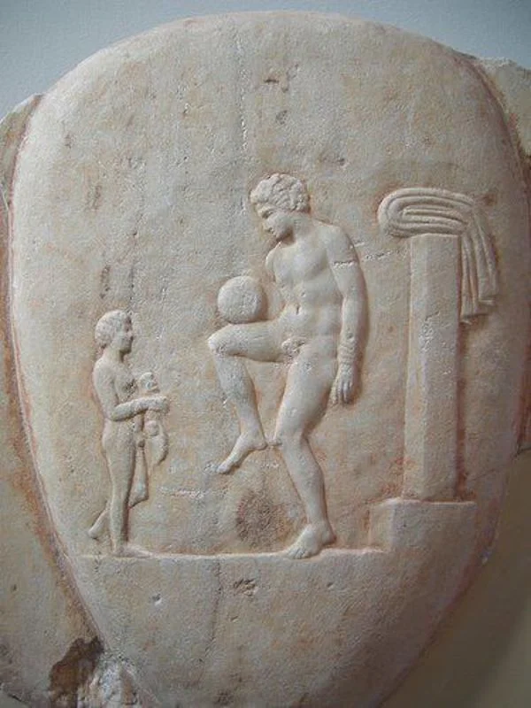 Ancient Greek football player balancing the ball. Part of a marble grave stele, found in Piraeus, 400-375 BC. The youth is watched by his young servant holding an aryballos and a strigil/National Archaeological Museum, Athens