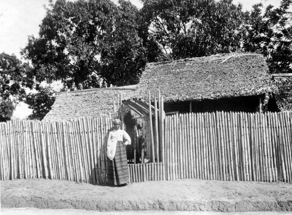 Betsinuisaraka people. Fenerive, Madagascar.  Typical fence with gate made of bamboo. The woman is the owner of the house, and is dressed in a skirt made of raffia fabric. Walter Kaudern's second expedition to Madagascar (1911-1912)/Wikimedia Commons 